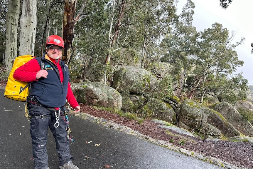 A man stands on a road with a pack on his back and boulders in the background. 