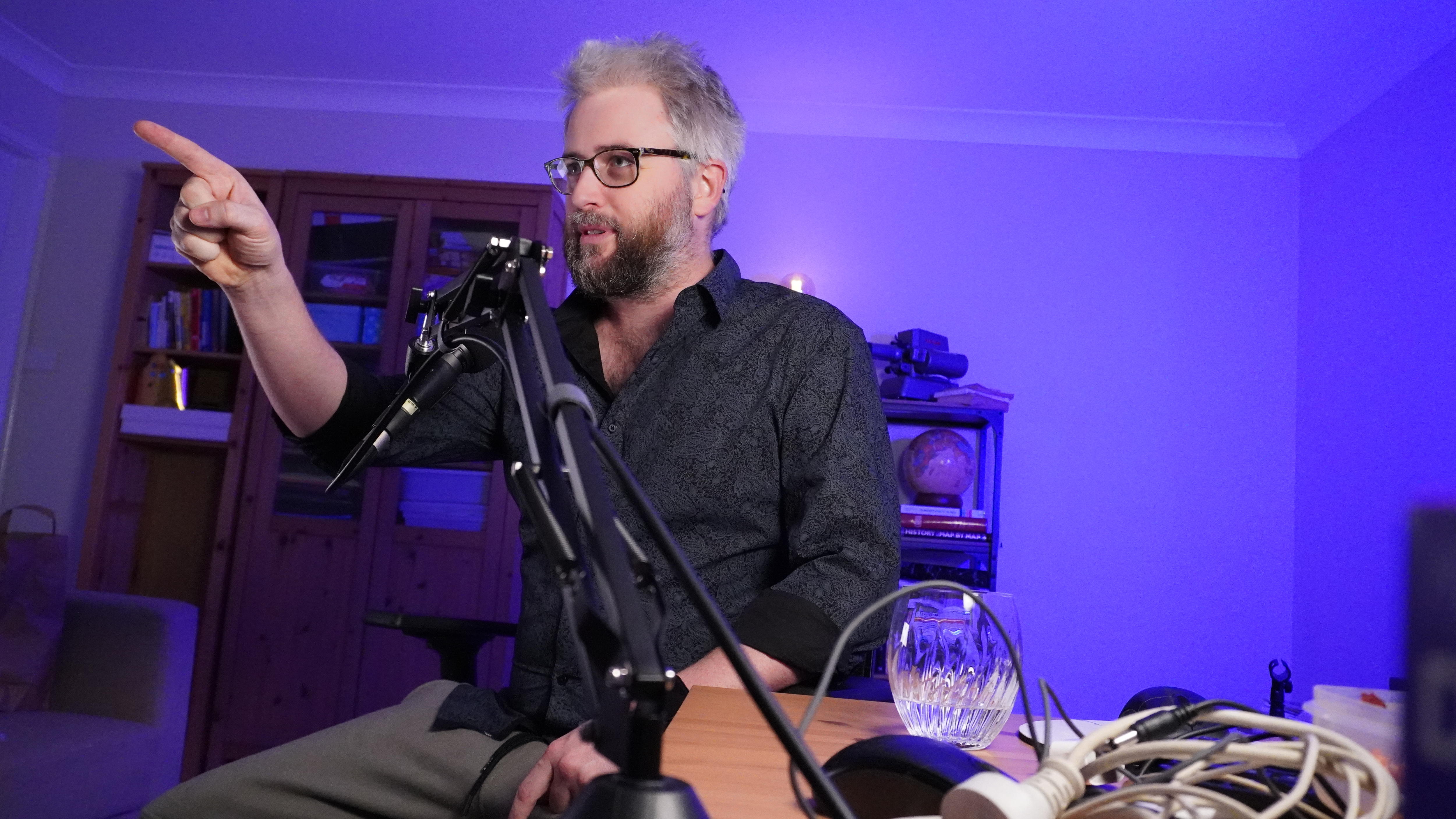 Man behind a microphone, pointing in front of himself, in a room with purple lighting.
