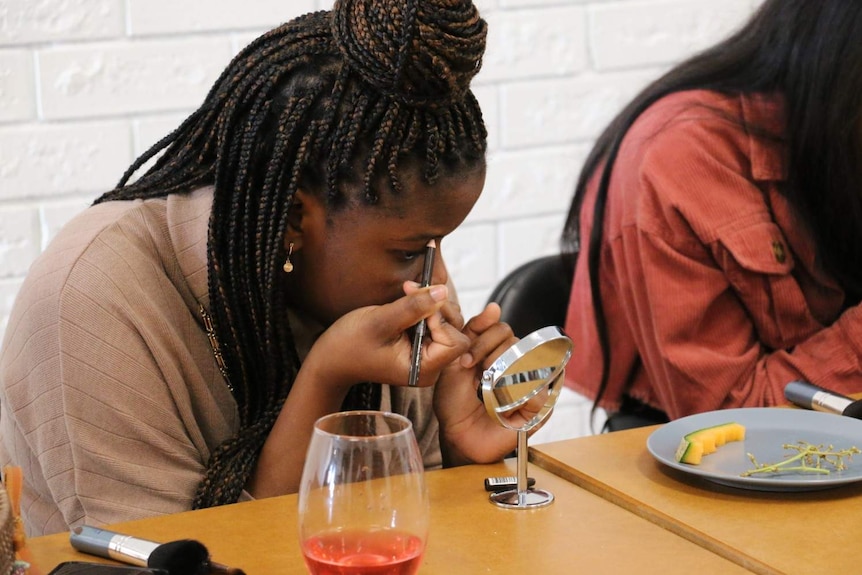 Milcent Tsvangirayi applies makeup at a table inside a hall.