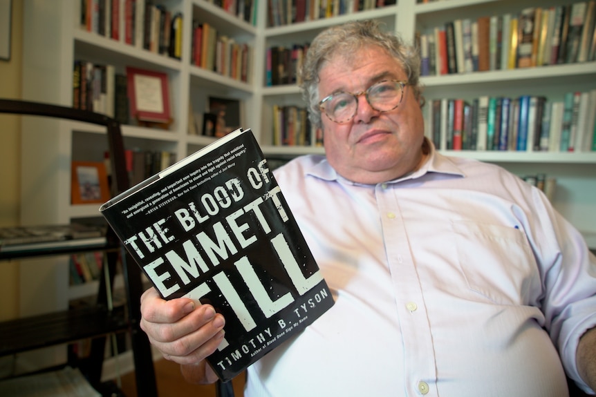 A man in a white button up top sitting in front of a wall of books holds up one titled The Blood of Emmett Till