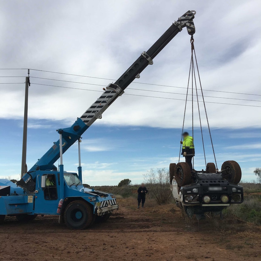 A crane lifts an upside down ute with a man in a fluoro jacket standing on the chassis.