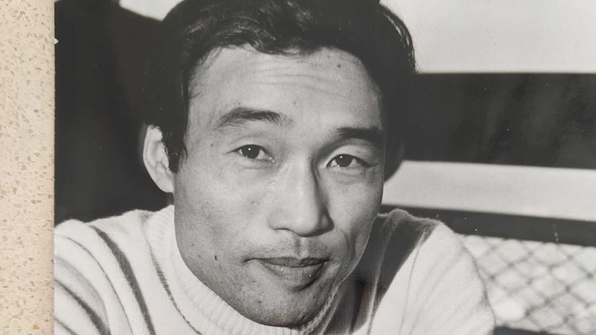 Black and white photograph of Kumi Taguchi's father, wearing white turtleneck leaning on table before him.