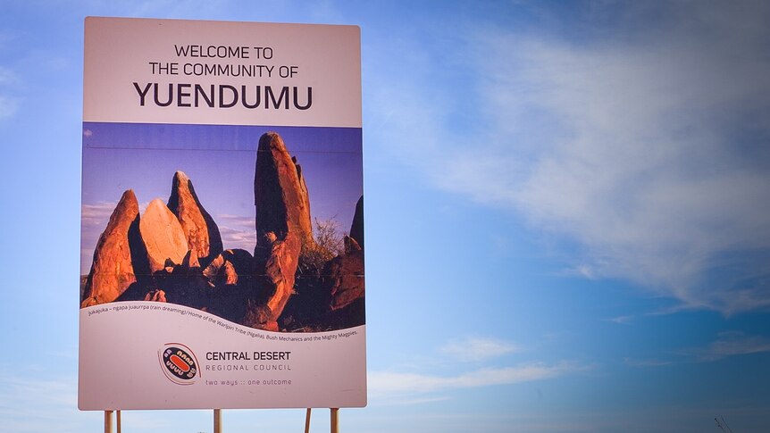 A sign reading 'welcome to the community of Yuendumu' and featuring a picture of red rocks, in front of blue sky