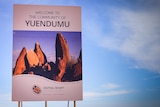 A sign reading 'welcome to the community of Yuendumu' and featuring a picture of red rocks, in front of blue sky