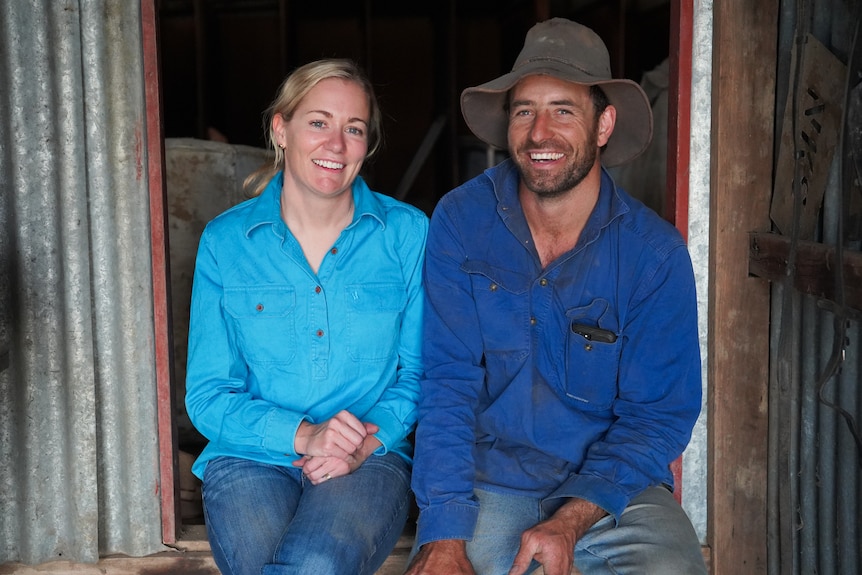 A woman in jade long sleeve and a man in long sleeve blue shirt, both smiling and sitting in front of a shed 