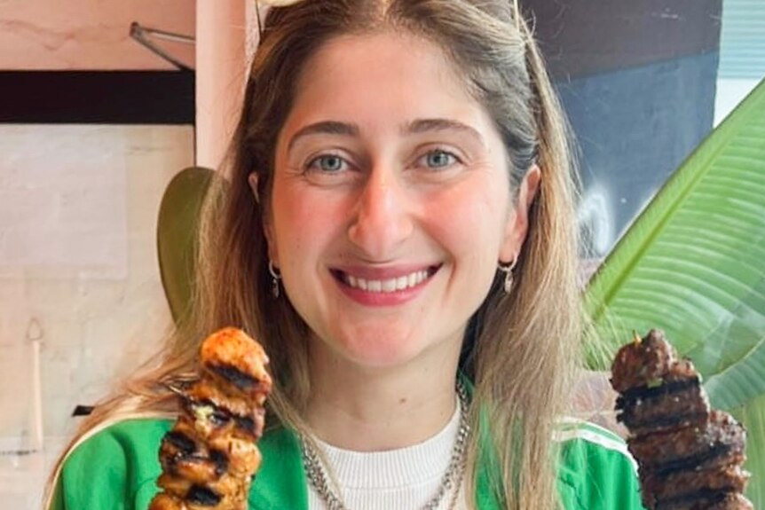 Kat is smiling at the camera while holding a souvlaki skewere of meat in each hand