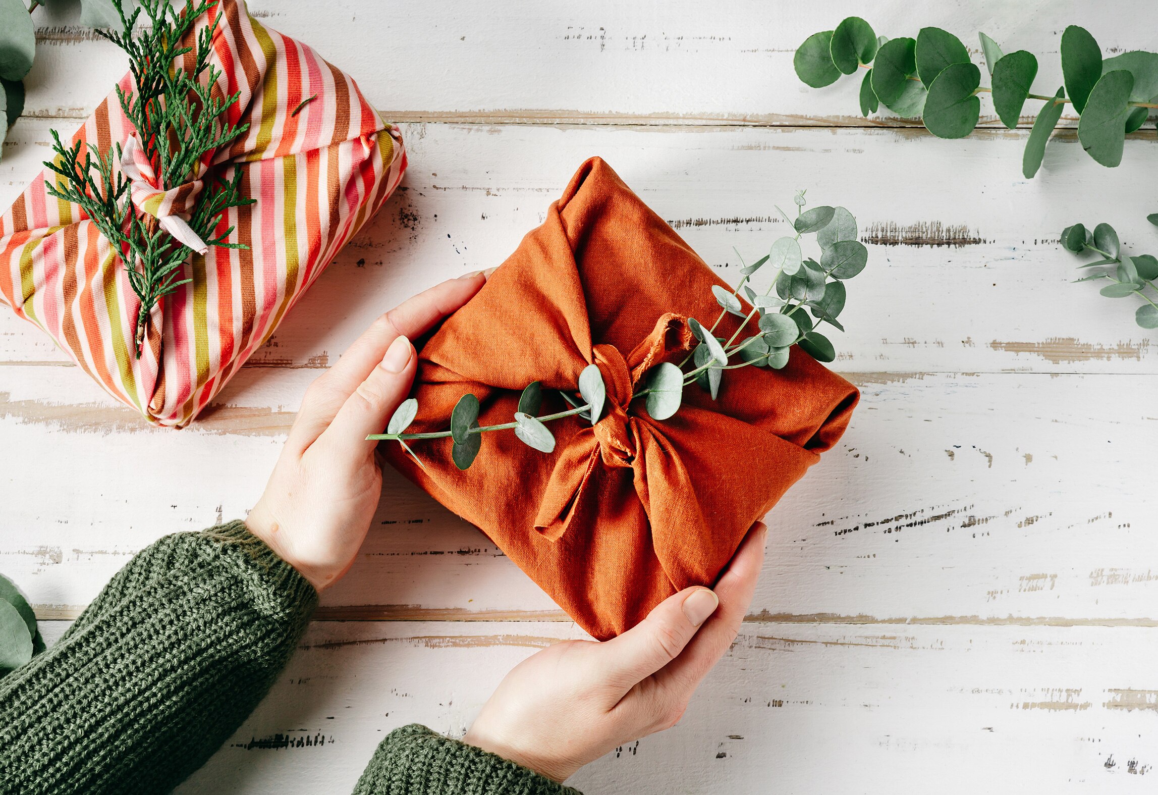 A woman's hands hold an orange furoshiki wrapped gift decorated with eucalyptus leaf stems on a white table.