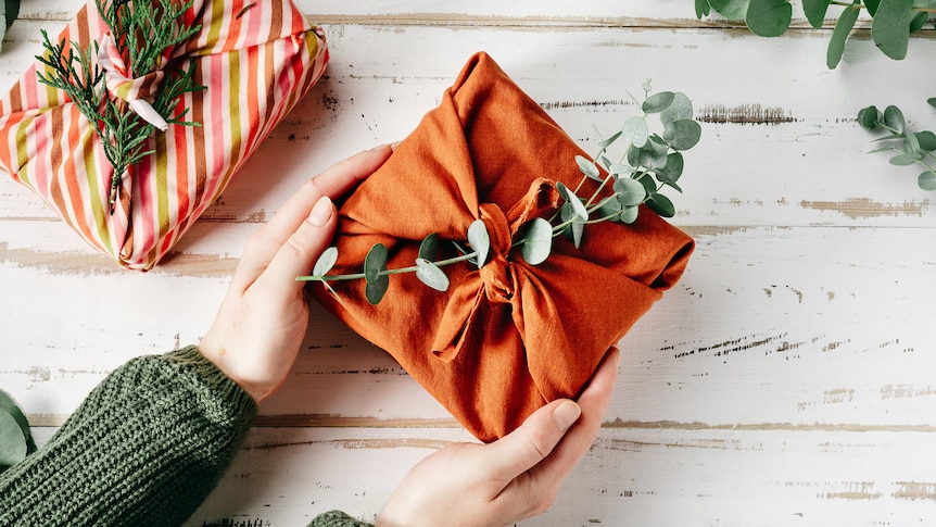 A woman's hands hold an orange furoshiki wrapped gift decorated with eucalyptus leaf stems on a white table.
