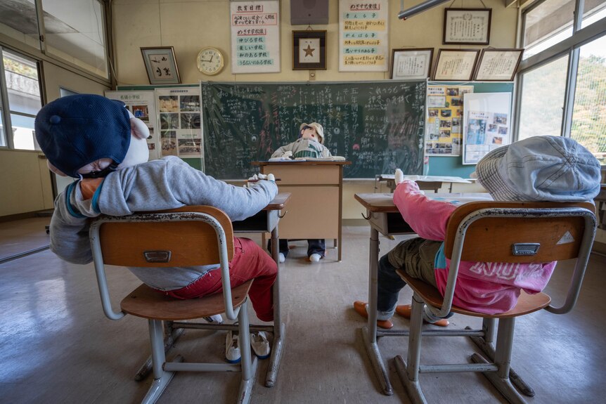Two dolls sit at desks facing another doll at the front of the classroom and a blackboard.