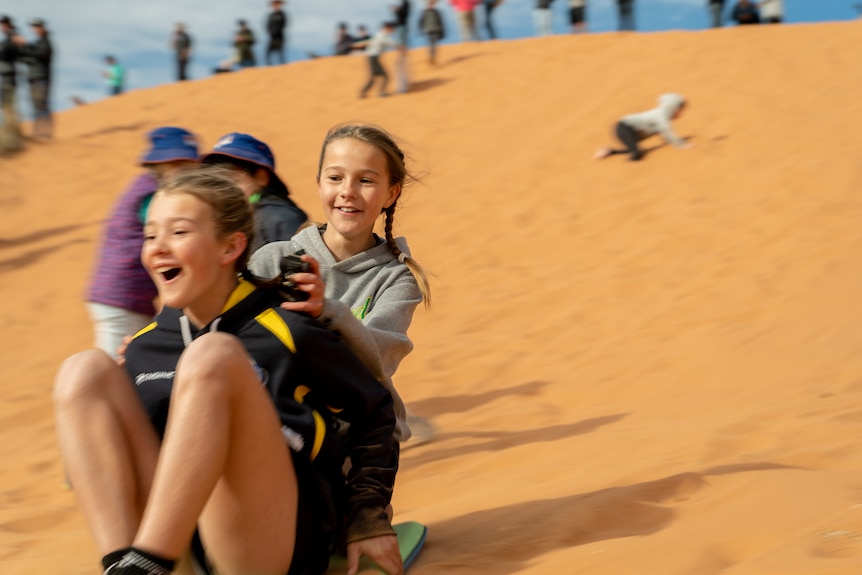 two girls on a board slide down a red sand dune while other children are climbing back to the top
