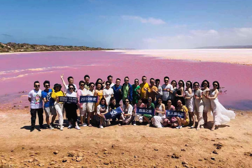 An Aus Highway Travel Services tour group poses for a photo standing in a big group in front of a pink lake.
