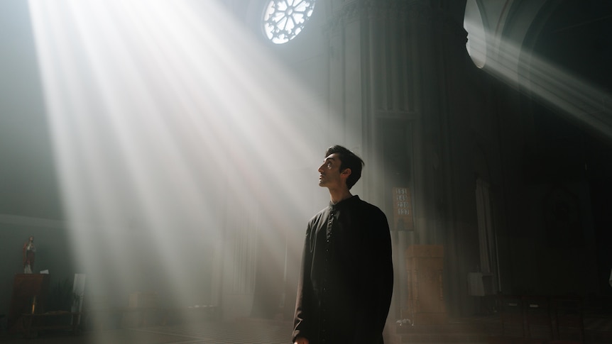 A lone priest stands in a beam of light in a church