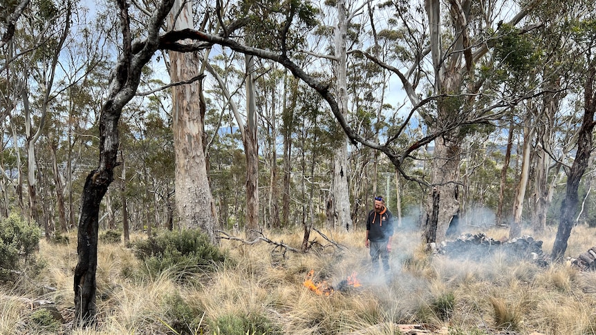 Jason Andrew Smith stands among trees with a small fire and smoke in front of him
