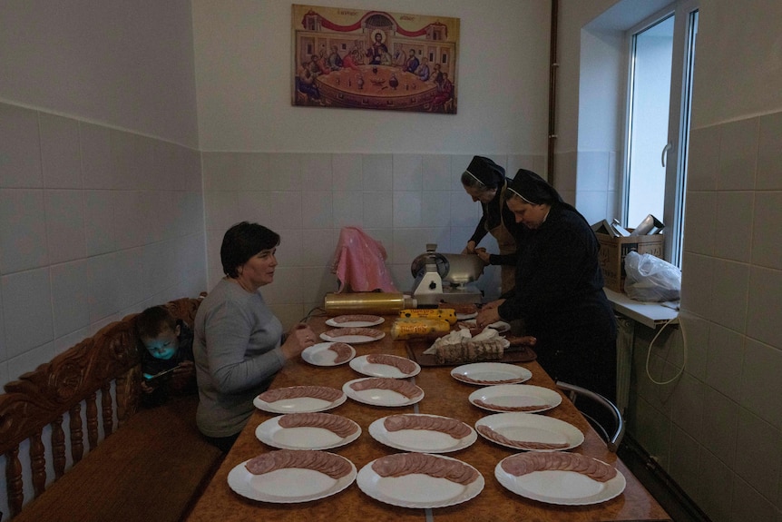 Two nuns stand cutting pieces of meat and placing on plates on a large table, as a woman and her son sit nearby.