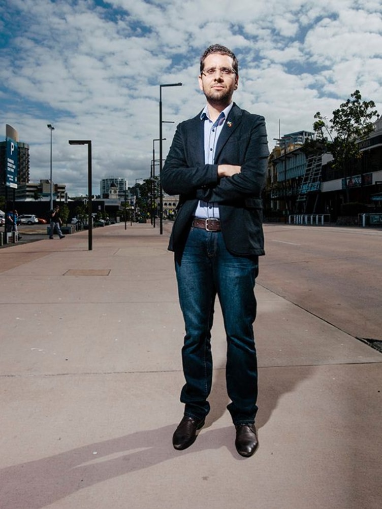 Man wearing jeans and a navy suit jacket stands with his arms crossed.