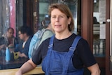A woman wearing a blue denim apron over a navy t-shirt stands in front of some cafe tables with patrons.