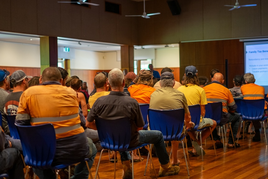 A group of people, mostly men and many in high-vis, sit in a community hall.