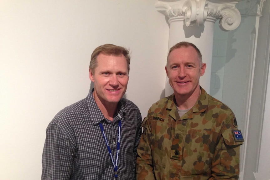 Two man stand in front of a white wall and look at the camera.