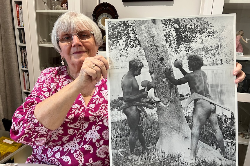 A woman in her 70s holding a black and white historical print of Aboriginal men cutting a shield from tree bark with stone axes