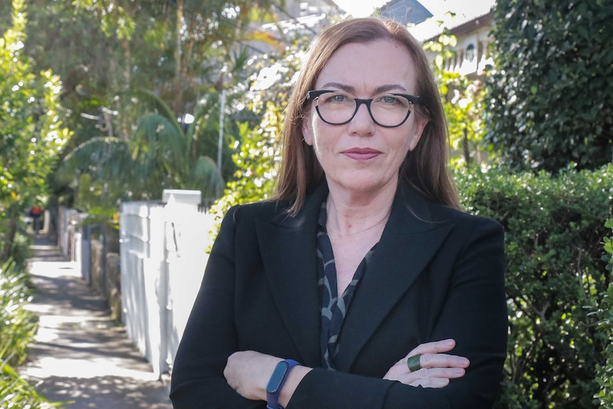 A woman in a black jacket looking at the camera, with arms crossed, on a suburban street.