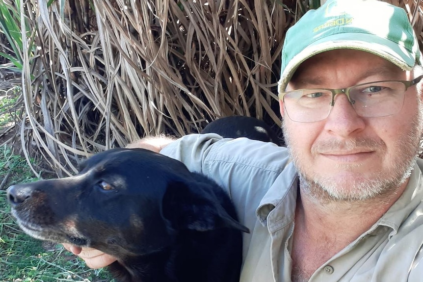 A man in a khaki collared shirt and a green baseball cap sits in front of a sugar cane crop with his dog.