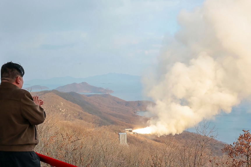 Kym Jong Un claps while watching a missile test in the distance along a rugged coastline