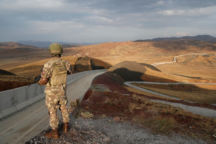 A soldier stands at the edge of a road overlooking rolling arid hills 