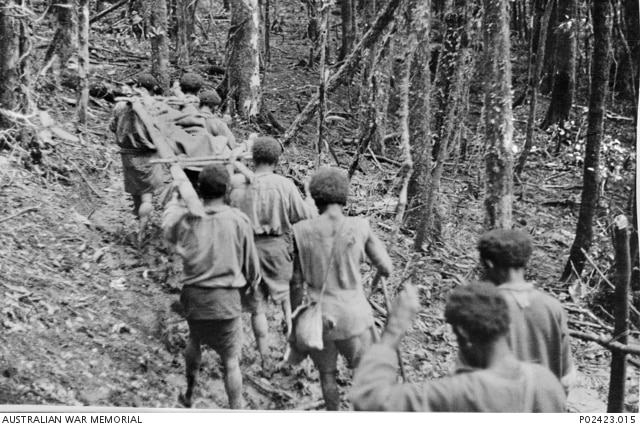 A black and white photograph shows a group of PNG men carrying a stretcher through the jungle