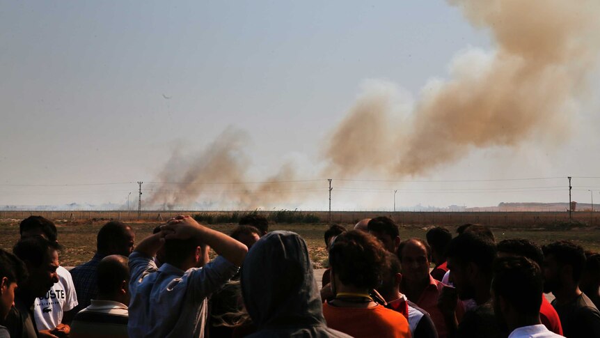 A group of people, some with hands on their heads, watch huge plumes of smoke billowing from military targets