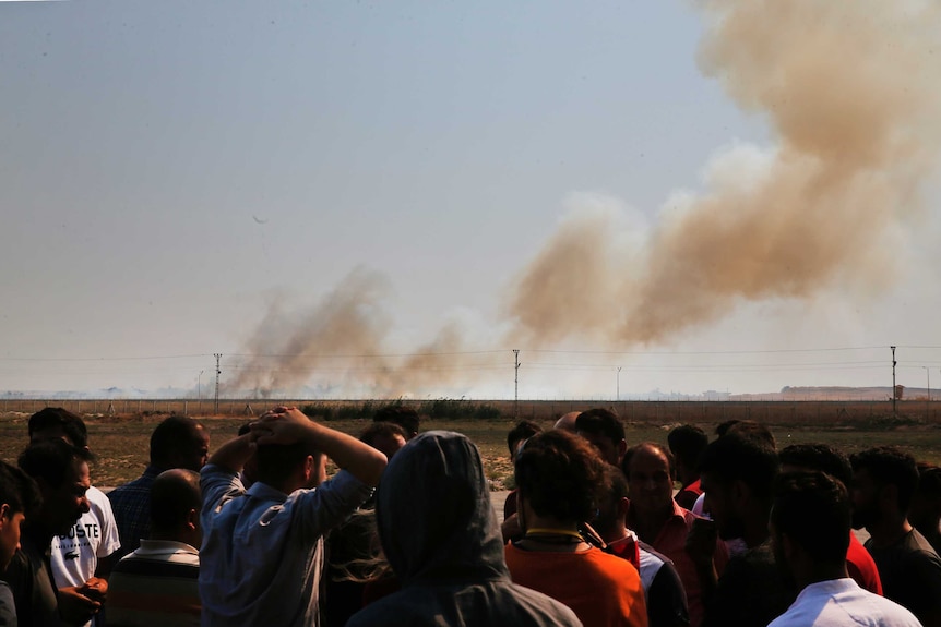 A group of people, some with hands on their heads, watch huge plumes of smoke billowing from military targets