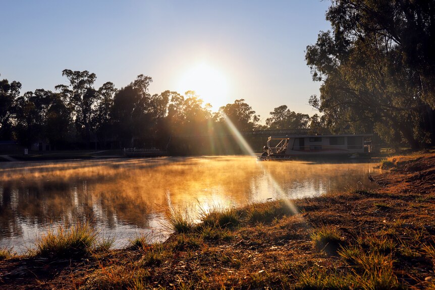 Early morning sunlight hits the Murray river with a bridge and house boat visible in distance