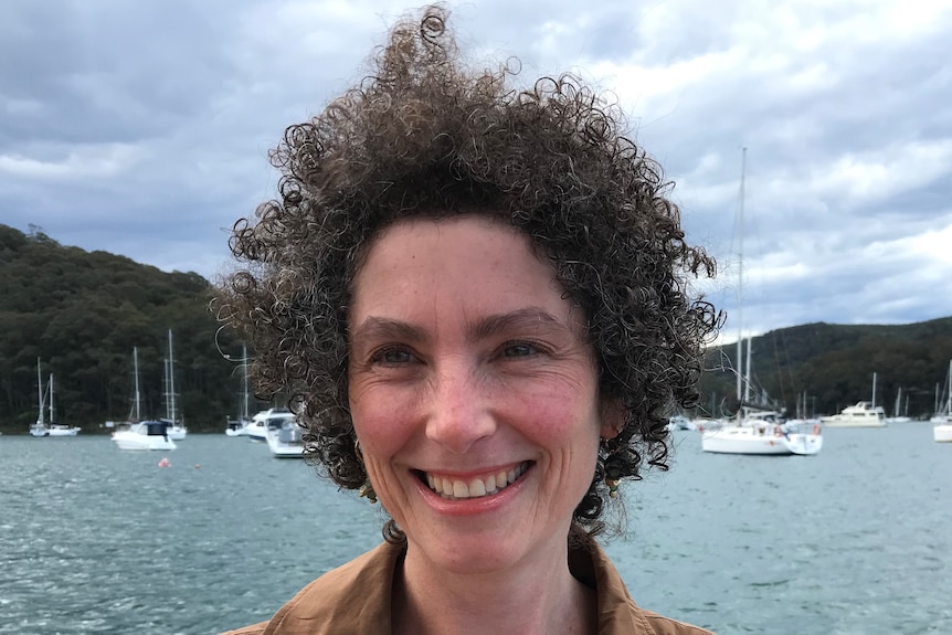 Headshot of a widely smiling woman with water and sailing boats in background. Curly hair afro, beige shirt visible.