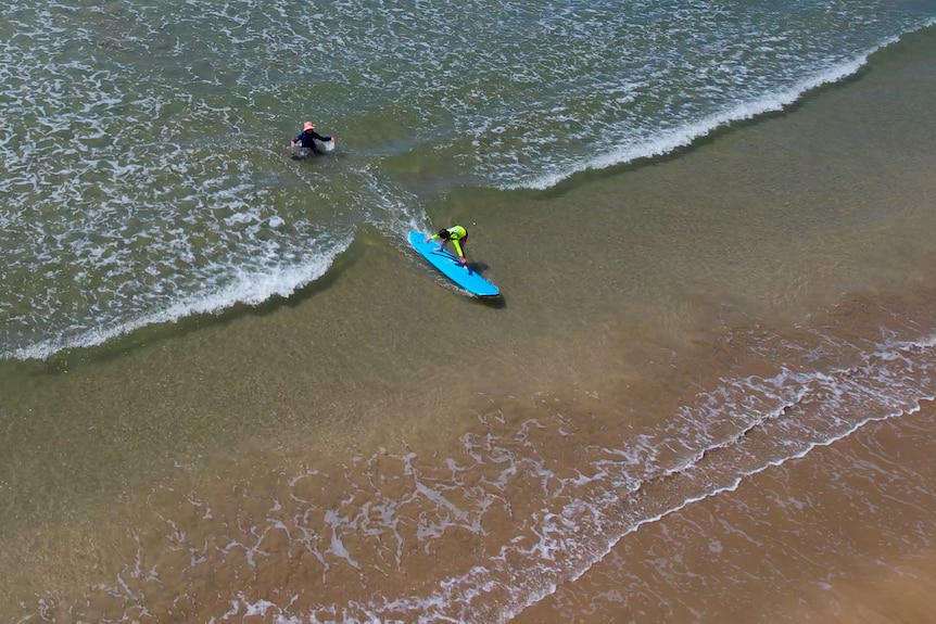 Drone image of girl standing up on a blue surfboard in clear water with woman behind