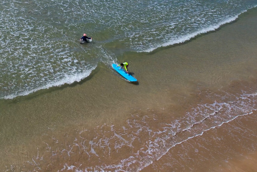 Imagen de un dron de una chica de pie sobre una tabla de surf azul en agua clara con una mujer detrás