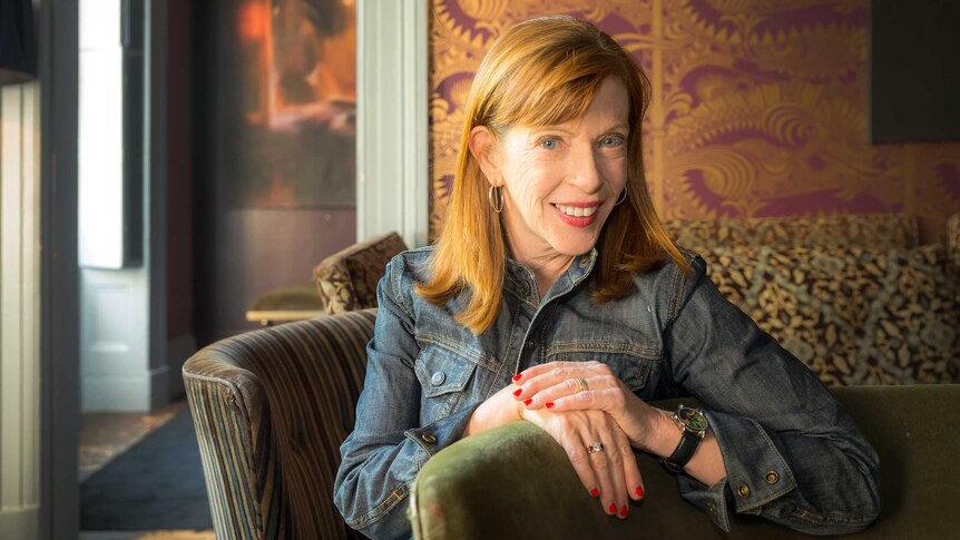A red-haired woman, author Susan Orlean, sits, smiling, on a chair in a living room with one hand lying on top of the other.