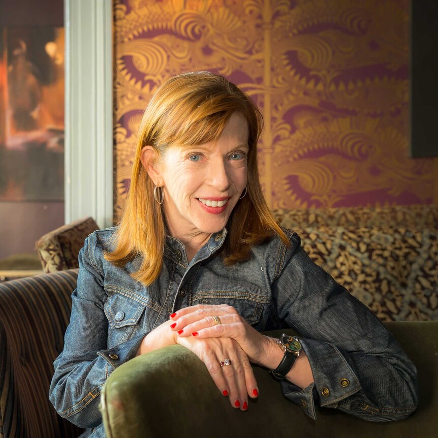 A red-haired woman, author Susan Orlean, sits, smiling, on a chair in a living room with one hand lying on top of the other.