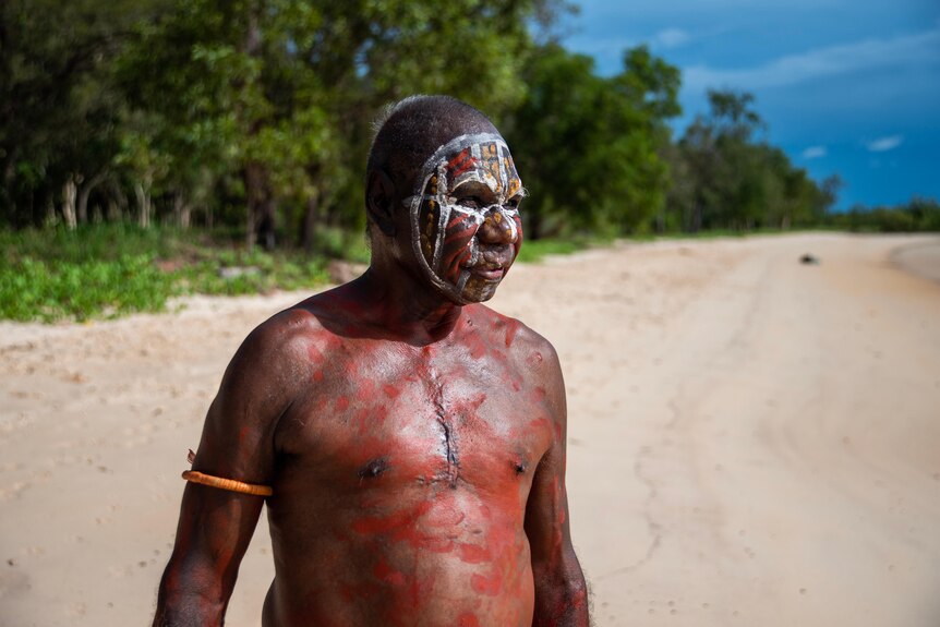 An Aboriginal man standing on a beach and looking serious, with his face and chest painted with traditional markings.