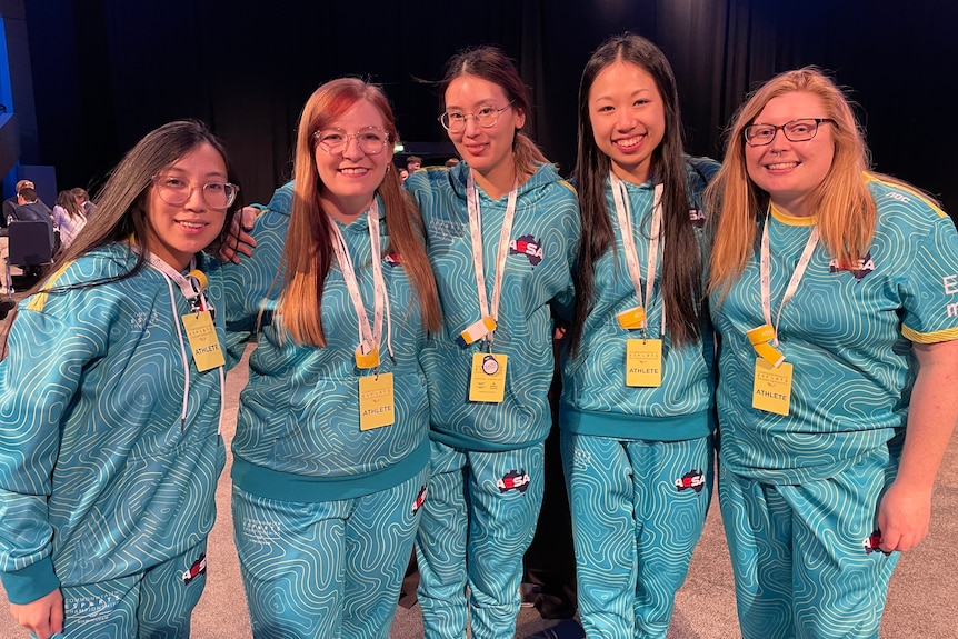 Five women wearing green and gold tracksuits stand arm in arm.