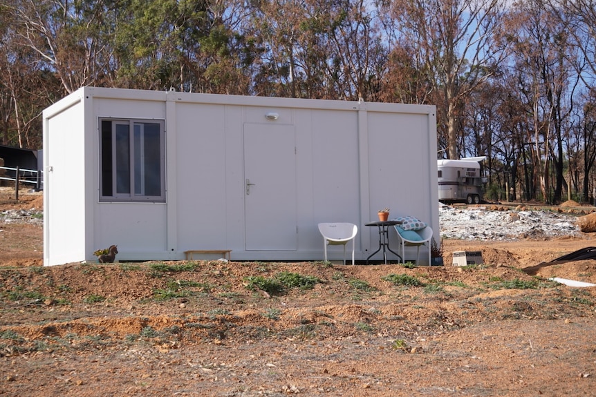 A white transportable shipping container-like building, with a window, and table and chairs out the front, on a rural property.