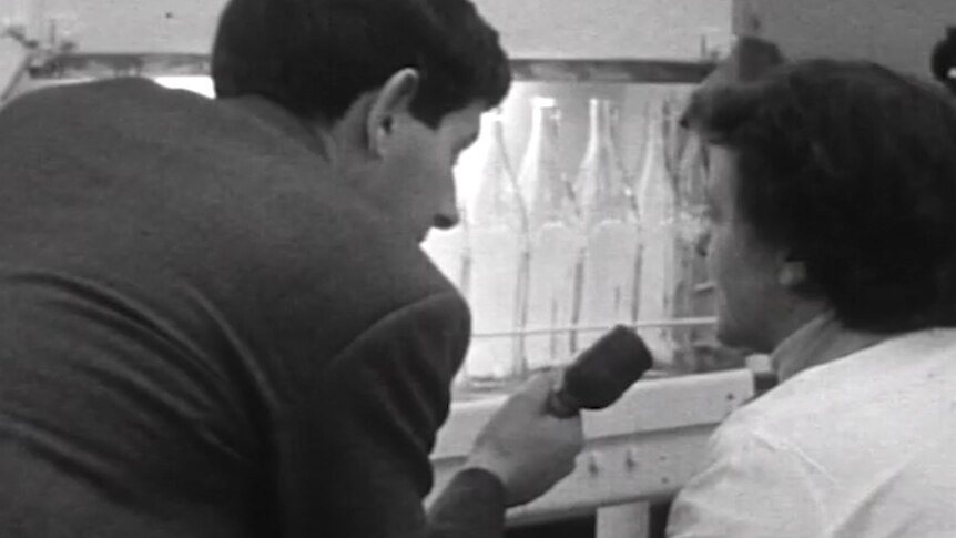 A man with a microphone talks to a woman sitting in a factory in front of a line of glass bottles on a conveyer belt