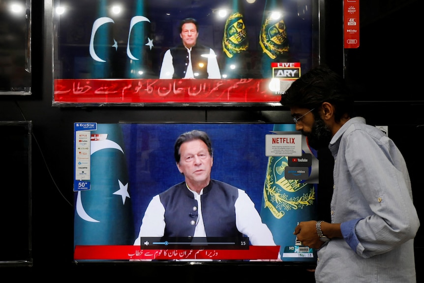 A South Asian shopkeeper in white shirt watches Imran Khan broadcasting on TV with Pakistan flags behind him