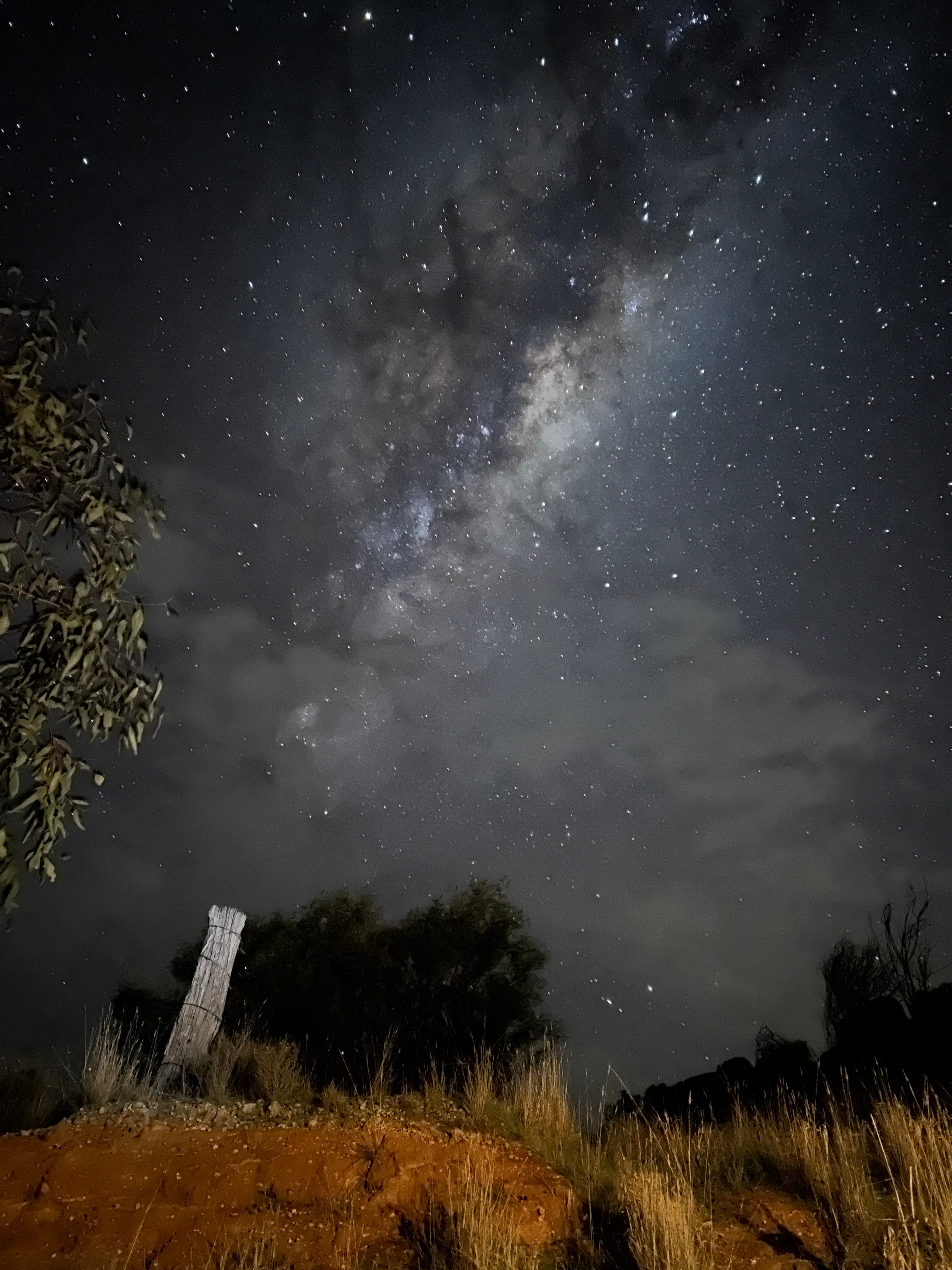 The Milky Way galaxy in the night sky, with a fence post, red dirt and dry grass illuminated in the foreground.