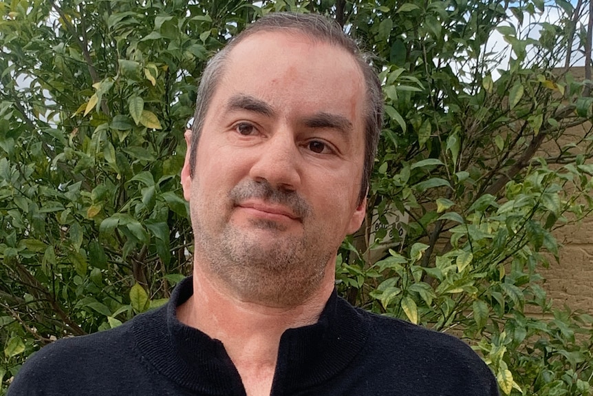 An unsmiling man wearing an open-necked shirt in a garden.