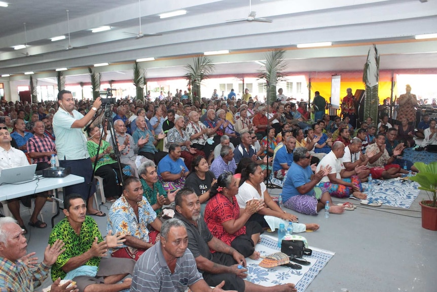 A huge, diverse crowd packs out a large function room, with people sitting on the ground and in rows of chairs.