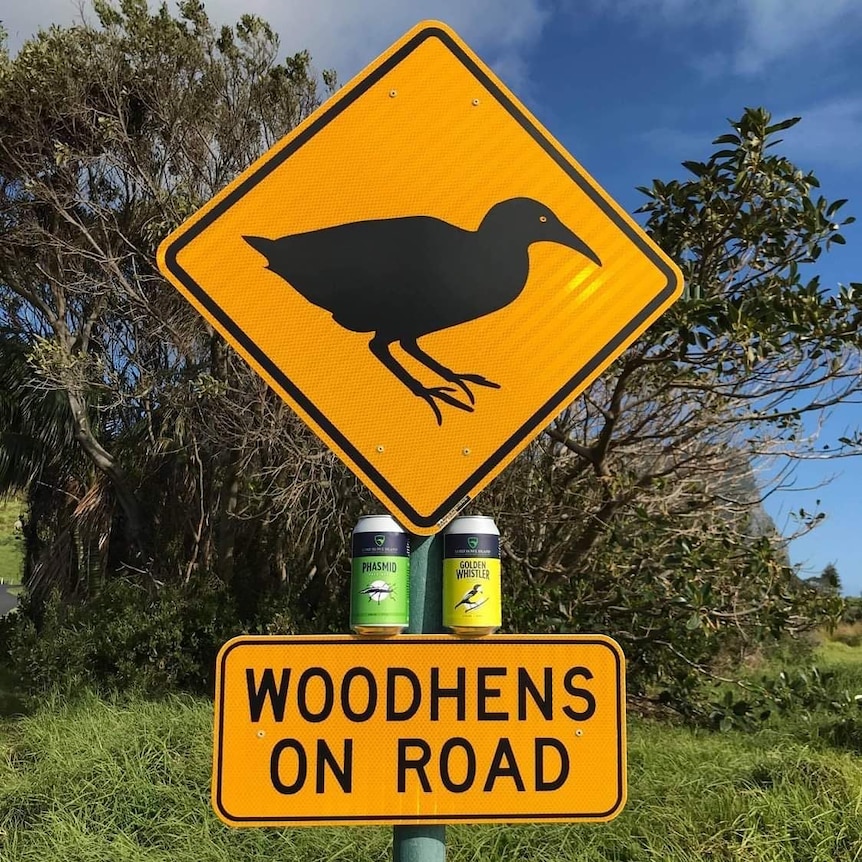 Two beers sit on a  road sign that reads 'Woodhens on road' and has the silhouette of a bird 