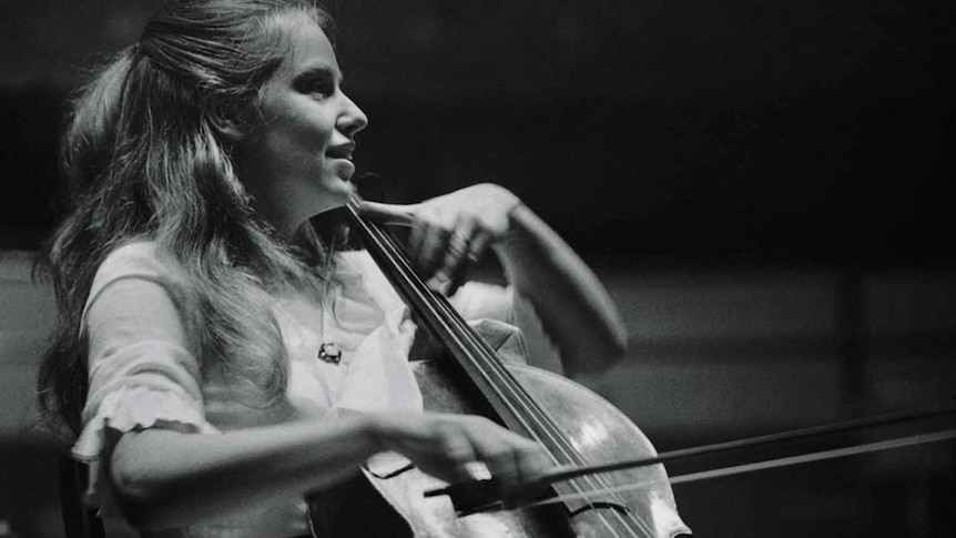 Black and white photo of cellist Jacqueline du pre smiling as she plays cello, looking to the right