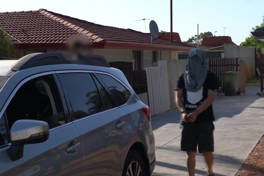 A man with a shirt over his head approaches a car in handcuffs