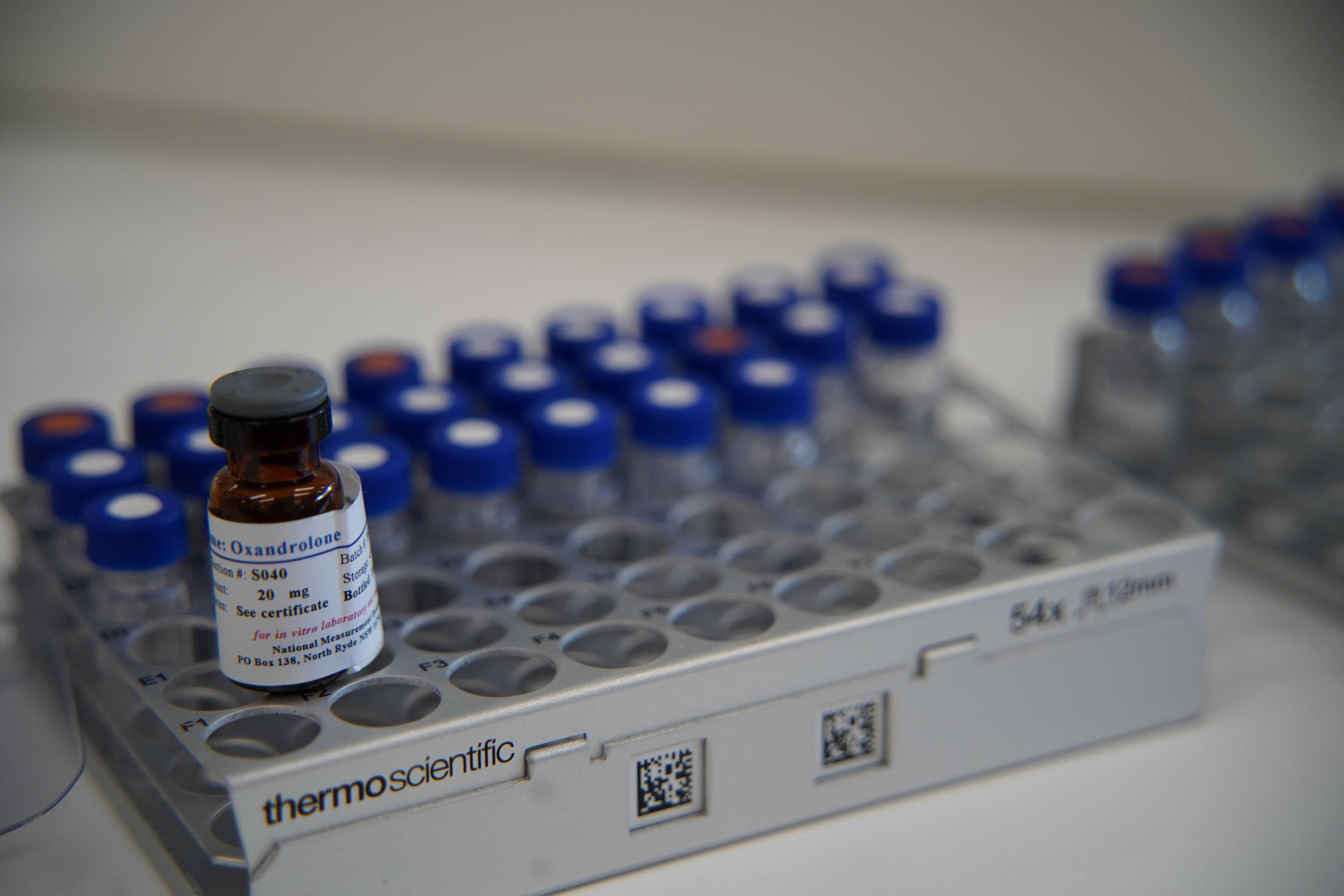 Um pequeno frasco marrom com rótulo branco está em foco com outros pequenos frascos ao fundo.