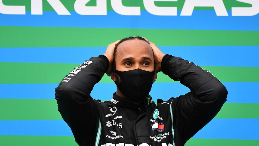 Lewis Hamilton stands with his hands on his head wearing a black face mask