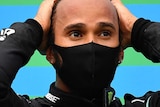 Lewis Hamilton stands with his hands on his head wearing a black face mask